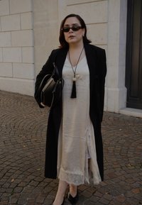 Cappotto nero sopra un vestito beige con orlo in pizzo. Gli accessori includono una borsa nera, una collana con pendente e frange, e occhiali da sole scuri.