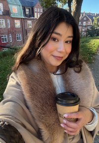 Beige kappa med en mjuk brun pälskrage, håller en svart kaffekopp med ärm i kraftpapper, mot en bakgrund av tegelbyggnader.