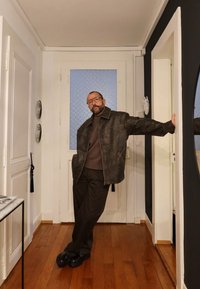 Brązowa skórzana kurtka o luźnym kroju, noszona na ciemnym swetrze i brązowych spodniach. Odbijające światło czarne buty. Neutralne tło wnętrza.