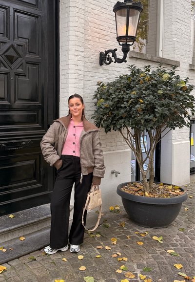 Mujer joven con chaqueta marrón, suéter rosa, pantalones negros y zapatillas, de pie en una calle empedrada junto a un árbol frondoso en una maceta grande.