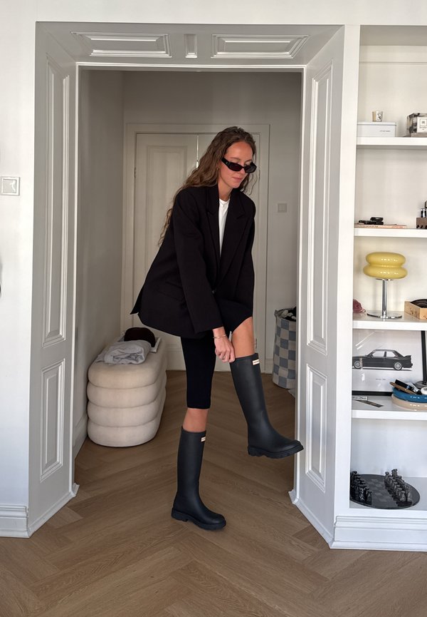 Una mujer con gafas de sol y ropa negra se prueba unas botas altas negras en una puerta. Cerca hay estanterías y un taburete acolchado.