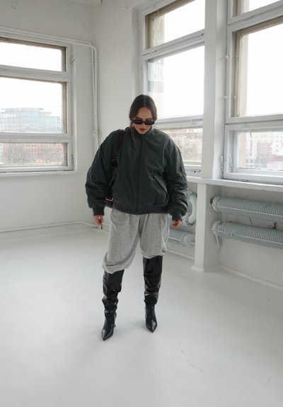 Mujer con chaqueta oscura y extragrande, pantalones grises sueltos, botas negras hasta la rodilla y gafas de sol dentro de una habitación luminosa con grandes ventanas.