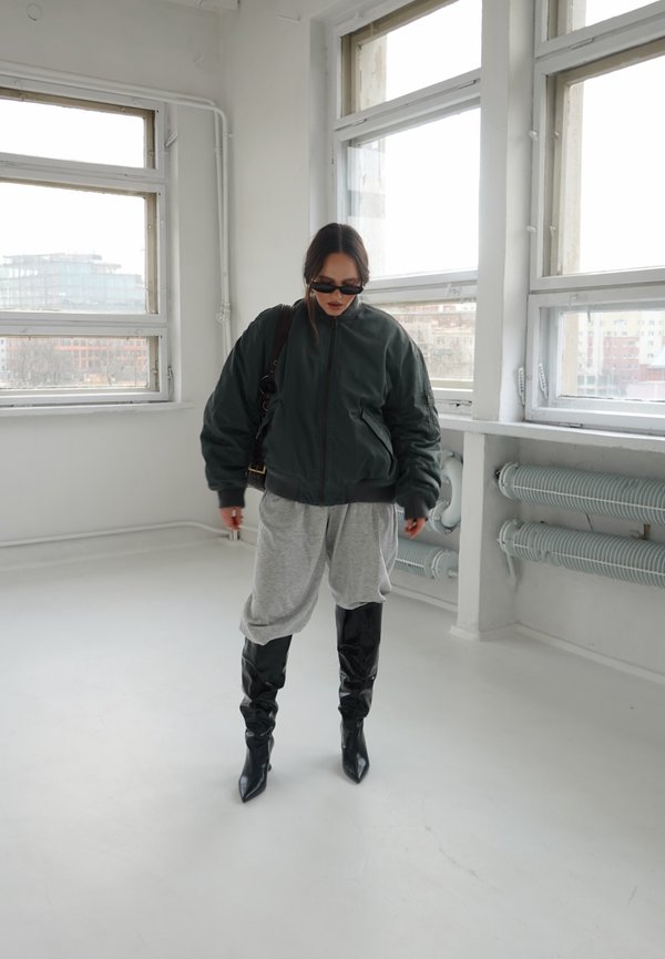 Žena ve tmavé nadměrné bundě, šedých volných kalhotách, černých kozačkách ke kolenům a slunečních brýlích v jasné místnosti s velkými okny.