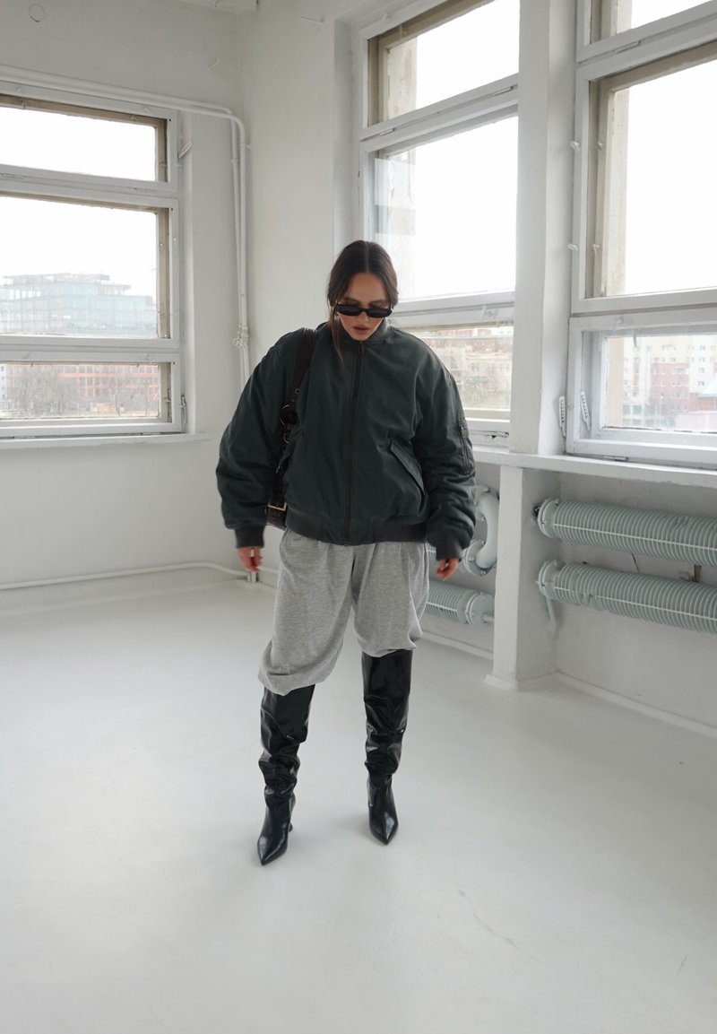 Γυναίκα που φοράει σκουρόχρωμο φαρδύ μπουφάν, γκρι χαλαρό παντελόνι, μαύρες μπότες μέχρι το γόνατο και γυαλιά ηλίου μέσα σε ένα φωτεινό δωμάτιο με μεγάλα παράθυρα.