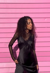 Femme aux cheveux bouclés portant un haut transparent à manches longues noir et une robe en satin, debout devant un mur à lattes horizontales rose vif.