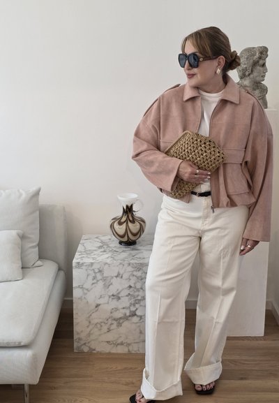 Mujer con chaqueta rosa empolvado de gran tamaño y pantalones crema sostiene un bolso tejido, de pie junto a un pedestal de mármol con jarrón y busto clásico.