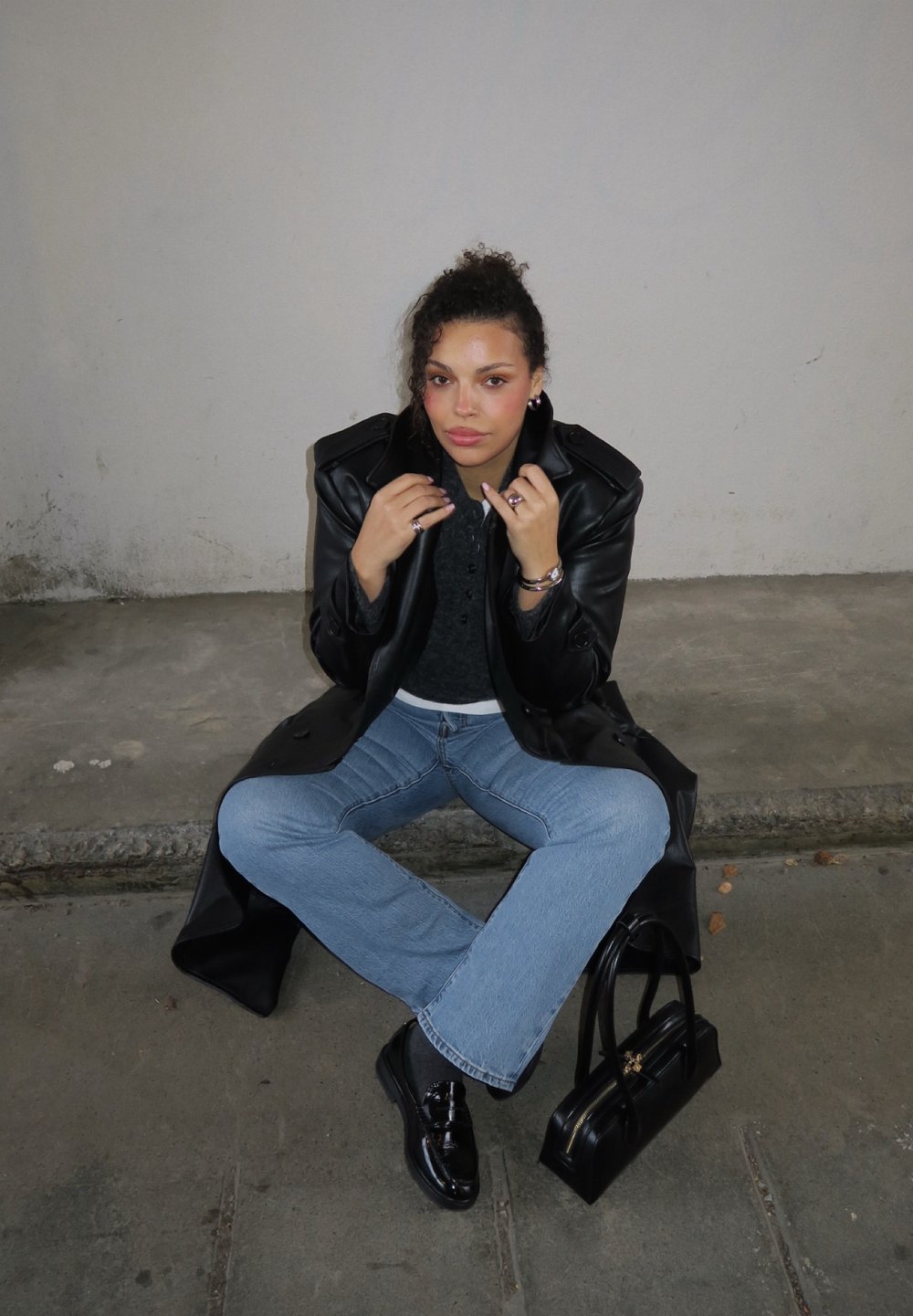 Sort læderfrakke over en grå strikket sweater, vidde blå jeans, skinnende sorte sko og en lille sort håndtaske. Siddende på en betonoverflade.