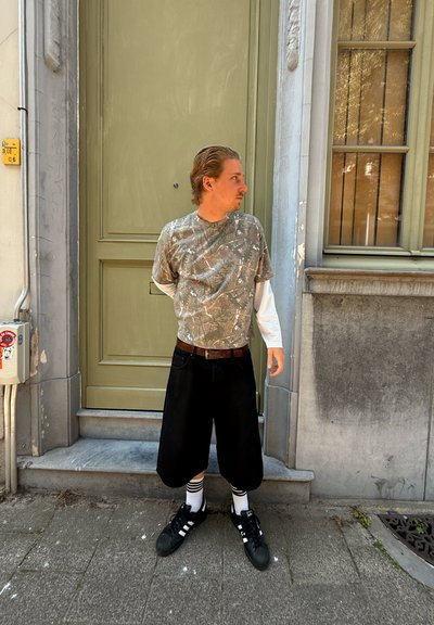 Conjunto casual que incluye una camiseta de patrón camuflaje, pantalones cortos anchos negros y zapatillas negras con rayas blancas. Cinturón marrón y calcetines blancos visibles.