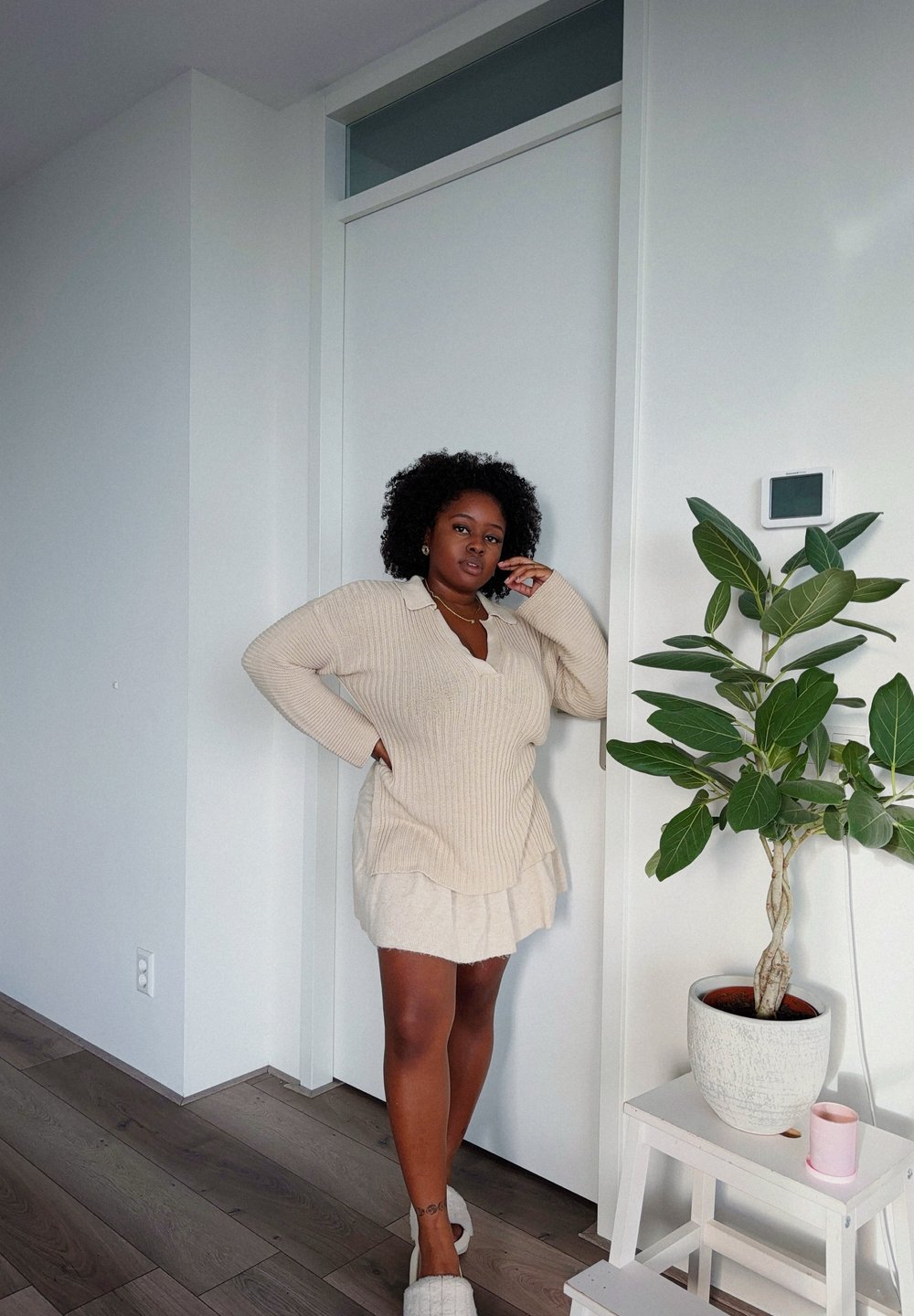 Beige oversized knitted sweater with a V-neck, layered over a cream skirt. Light wooden floor, green plant in a white pot, and a wall-mounted device visible.