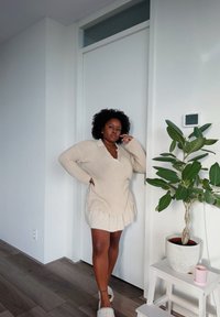 Beige oversized knitted sweater with a V-neck, layered over a cream skirt. Light wooden floor, green plant in a white pot, and a wall-mounted device visible.
