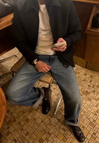 Melns bumbvedēja stila džemperis pār baltu t-kreklu, zilas džinsu bikses ar apkaklēm un brūnas kurpes, sēžot uz austas krēsla pret rakstainu grīdu.