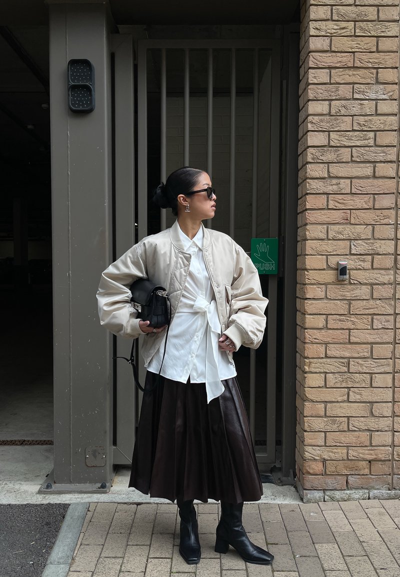 Frau mit Sonnenbrille, die eine beige Jacke, ein weißes gebundenes Hemd, einen schwarzen plissierten Rock und schwarze Stiefel trägt, steht bei einem eingezäunten Eingang mit Ziegelmauer.