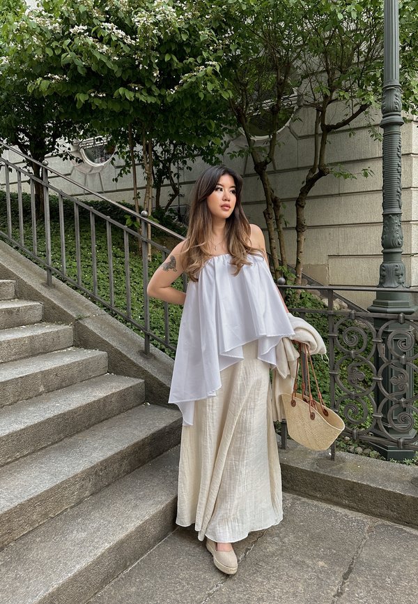 Weißes, schulterfreies Oberteil mit mehrlagigem Design; hellbeige, strukturierten Maxirock; beigen Espadrilles; gewebte Tasche mit Lederakzenten. Steinstufen und Grünpflanzen.