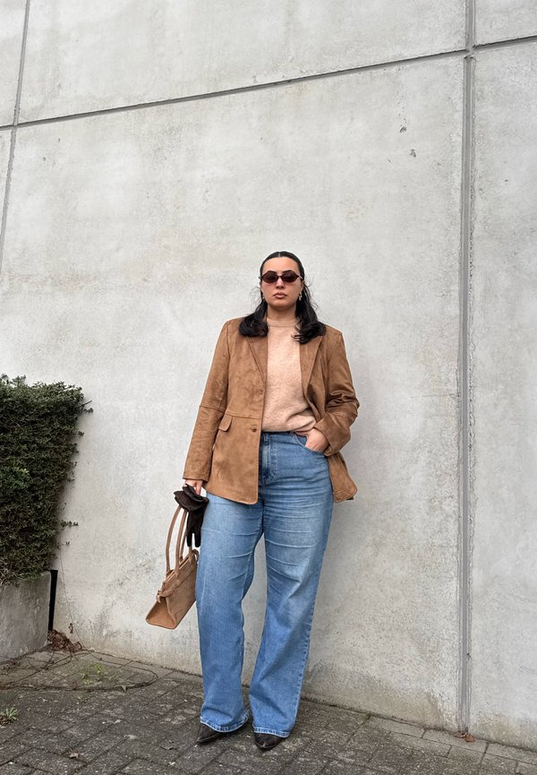 Femme portant des lunettes de soleil, un blazer marron, un pull beige, un jean bleu, tenant un sac à main marron et des gants, debout contre un mur en béton.