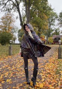 Poncho impermeabile a quadri in verde scuro e marrone, con cappuccio e chiusura frontale. Indossato sopra leggings neri e stivali di gomma neri. Sfondo di fogliame autunnale.