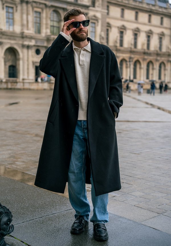 Man wearing a black long coat, white jumper, blue jeans, and sunglasses stands on a wet street in front of a historic building.