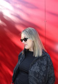 Kobieta z blond włosami, nosząca czarne okulary przeciwsłoneczne i ciemną, wzorzystą kurtkę, stoi na tle jasnoczerwonej ściany z cieniami.