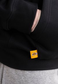 Zwarte capuchontrui met een textuurstof, voorzien van een gele en paarse Los Angeles Lakers-logo label nabij de zak. Grijze broek zichtbaar onderaan.