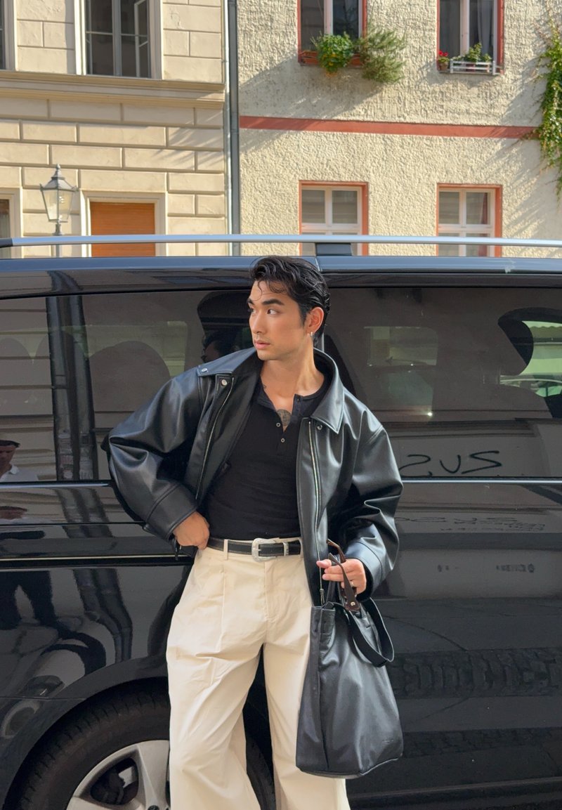 Person mit schwarzer Lederjacke und schwarzem Shirt, beigen Hosen, der eine schwarze Tasche hält, steht neben einem schwarzen Lieferwagen in einer städtischen Umgebung mit Gebäuden im Hintergrund.