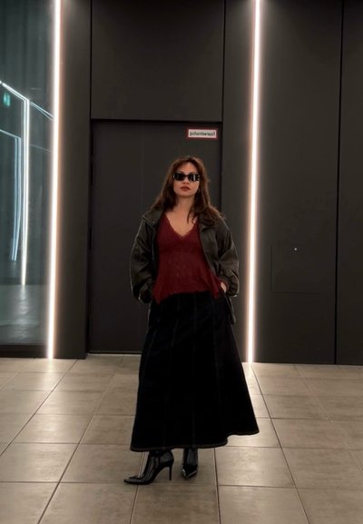 Mujer con gafas de sol, chaqueta oscura, camiseta roja, falda larga negra y botas negras con tacón, de pie en un pasillo moderno con tiras de luz verticales.