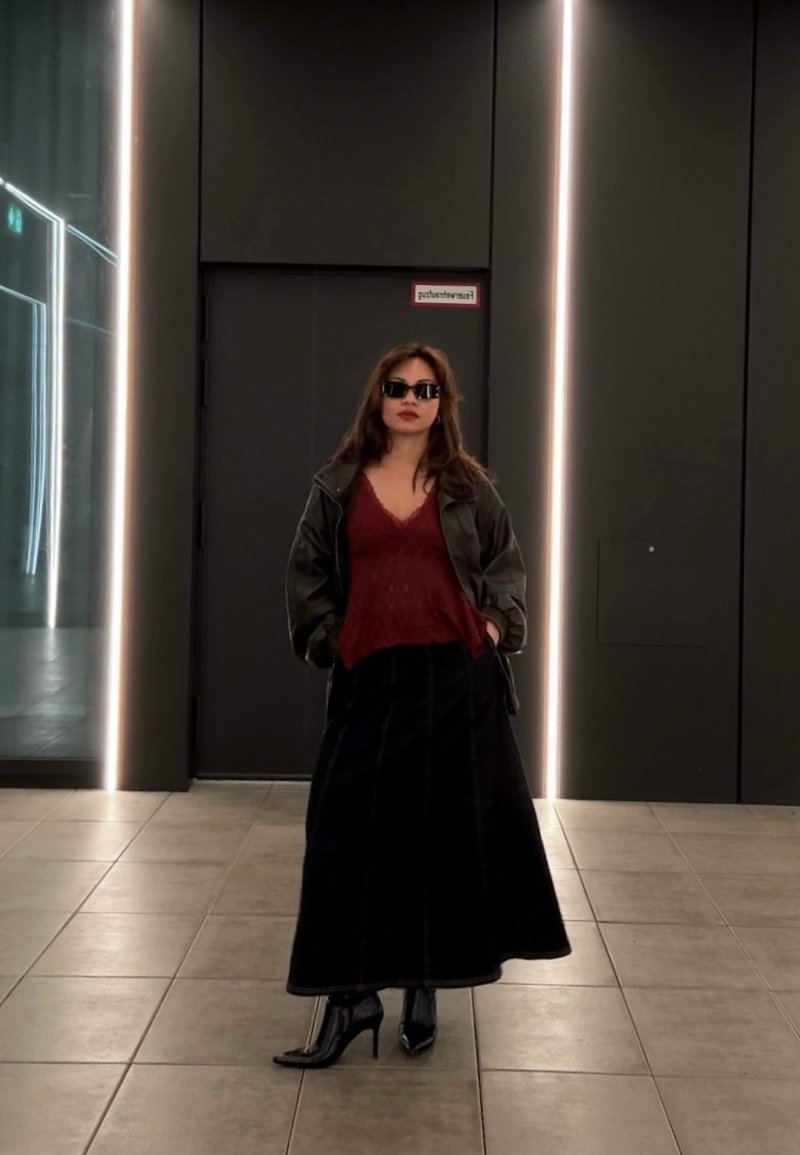 Femme portant des lunettes de soleil, une veste sombre, un haut rouge, une longue jupe noire et des bottes noires à talons, debout dans un couloir moderne avec des bandes lumineuses verticales.