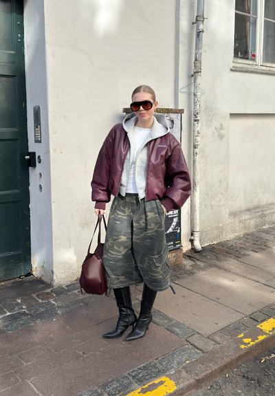 Mujer con chaqueta de cuero burdeos extragrande, sudadera gris con capucha, pantalones anchos militares, botas negras, gafas de sol grandes, sosteniendo bolso burdeos en la acera.