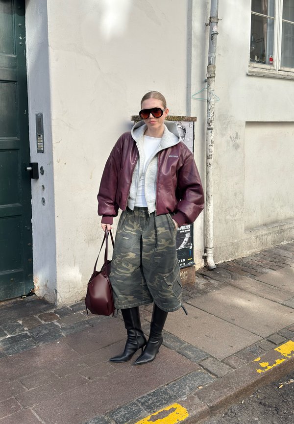 Ženska, ki nosi preveliko bordo usnjeno jakno, sivo kapucarico, široke maskirne hlače, črne škornje, velike sončna očala, drži bordo torbico na tlakovanju.