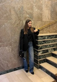 Czarna marynarka na czarnym topie, w połączeniu z szerokimi niebieskimi dżinsami. W ręku małą torebkę. Stoi na tle marmurowej ściany ze złotymi akcentami.