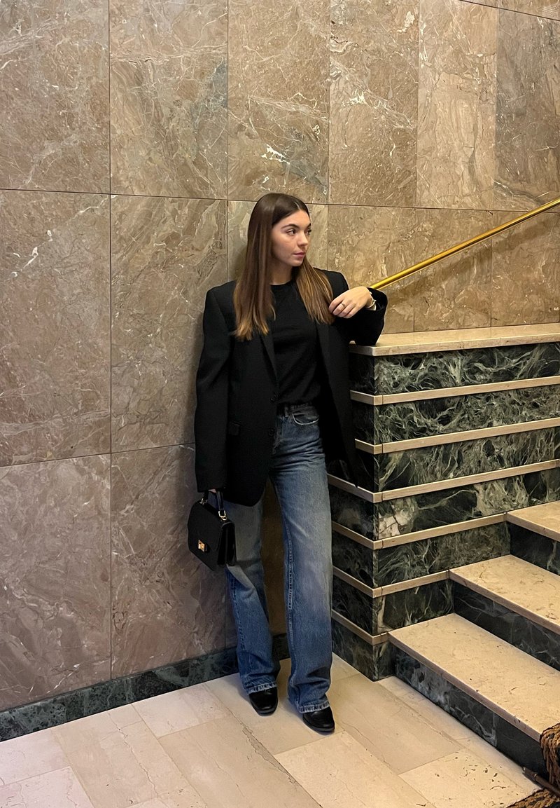 Sacou negru peste un top negru, asortat cu blugi largi, albaștri. Poartă o geantă mică în mână. Stă în picioare în fața unui perete de marmură cu accente aurii.