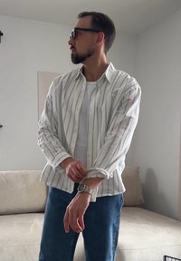 Άνδρας με γένια και γυαλιά που σηκώνει τα μανίκια ενός λευκού ριγέ πουκάμισου πάνω από λευκή μπλούζα, στέκεται σε ένα σαλόνι με ουδέτερους τόνους.