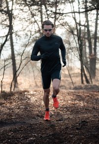 Athlète courant sur un sentier, portant un haut à manches longues noir et un short noir avec des accents blancs, ainsi que des chaussures de course orange vif.