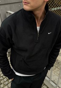 Sweat-shirt noir Nike zippé avec un col haut, fabriqué dans un matériau doux, avec un petit logo blanc sur la poitrine et deux poches latérales.