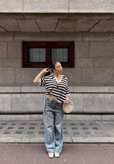 Jersey a rayas en blanco y negro de estilo crochet, jeans de pierna ancha azul claro y un bolso de mimbre. Calle suave con un fondo de pared de piedra texturizada.