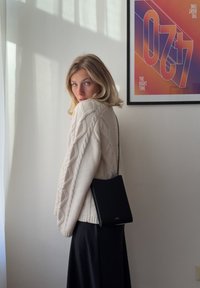 Jauna moteris, vilkinti grietinėlės spalvos mezginį ir juodą sijoną, stovi prie baltos sienos su spalvotais rėminiais plakatais, nešdama mažą juodą petinę rankinę.