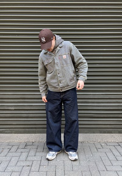 Chaqueta de mezclilla a rayas, sudadera gris por debajo, jeans de pierna ancha oscuros, zapatillas gris y blancas, y una gorra marrón con logo blanco contra un fondo texturizado.