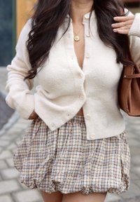 Cárdigan de punto beige con botones, combinado con una minifalda de cuadros marrones con un diseño recogido. Sin patrones prominentes ni detalles de textura.