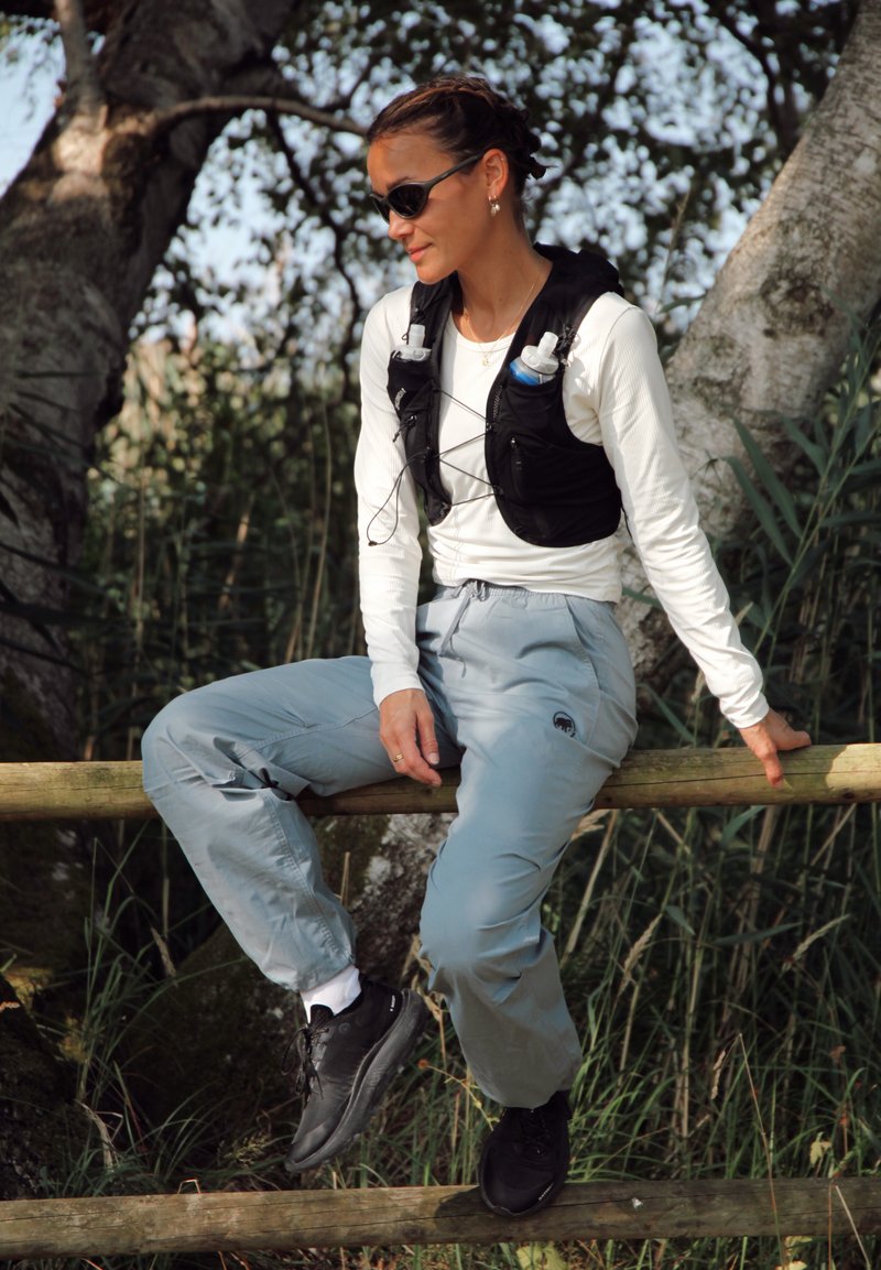 Donna con occhiali da sole, camicia bianca a maniche lunghe, pantaloni grigi e gilet nero con bottiglie d'acqua seduta su una recinzione di legno in un'area boschiva all'aperto.