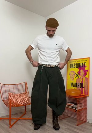 Hombre con boina marrón, camiseta blanca, pantalones anchos negros y botas, de pie en una habitación con una silla de alambre naranja y un póster de Basquiat Warhol.
