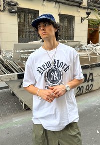 Balta per didelė T-marškiniai su juodu „New York“ grafiniu atvaizdu ir emblemą, alyvuogių žalios spalvos baggy šortai, sidabriniai papuošalai ir tamsiai mėlynas raštuotas kepuraitė.