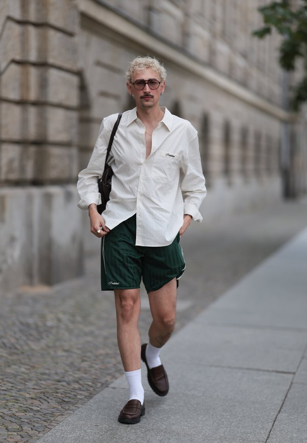 Ung mand med krøllet blond hår iført en hvid skjorte, mørkegrønne shorts, hvide sokker og brune sko går på en byfortov.