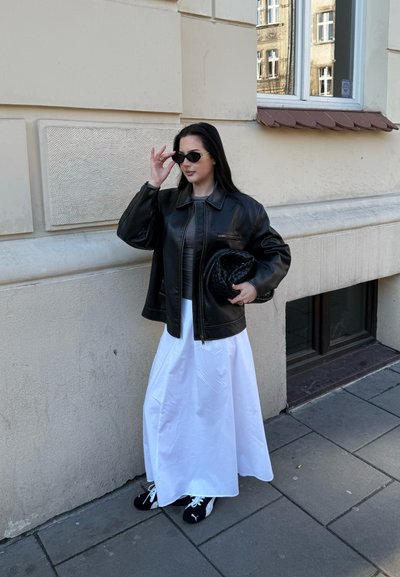 Chaqueta de cuero negra, vestido maxi blanco, zapatillas negras y gafas de sol. Pared texturizada y ventana de fondo.