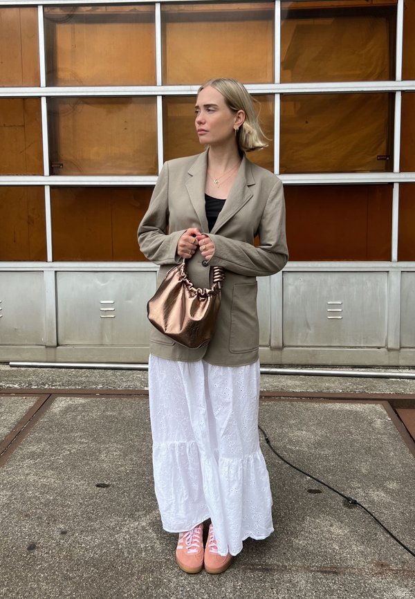 Beige blazer, zwarte top, witte maxi rok met oogjes, roze sneakers en een metalen bronzen handtas. Model staat tegen een industrieel decor.