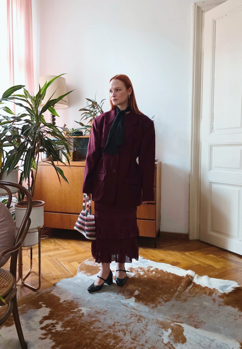 A woman wearing an oversized maroon blazer and a matching skirt stands on a cowhide rug in a living room, holding a striped beaded bag.