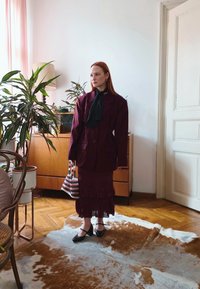 A woman wearing an oversized maroon blazer and a matching skirt stands on a cowhide rug in a living room, holding a striped beaded bag.