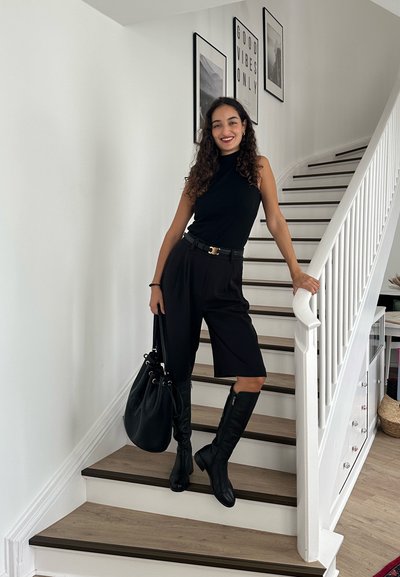 Top negro sin mangas, pantalones negros de cintura alta, botas negras hasta la rodilla. Sostiene un bolso negro. Escaleras de madera y obras de arte enmarcadas en el fondo.