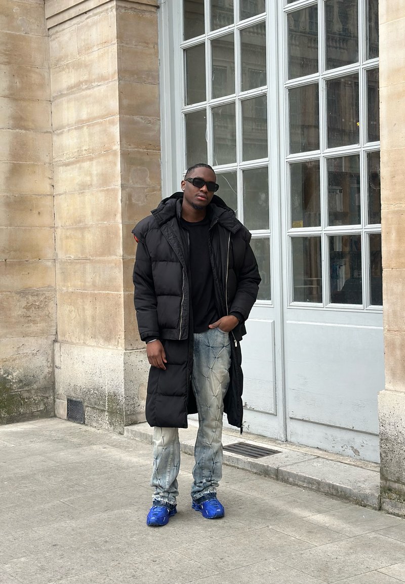 Homme portant un long manteau matelassé noir, une chemise noire, un jean tie-dye et des baskets bleues, debout près d'une grande porte vitrée sur un sol en pierre.