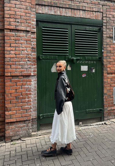 Chaqueta de cuero negra corta sobre una falda blanca y fluida. La modelo lleva zapatos de plataforma negros con cordones y lleva un bolso brillante.