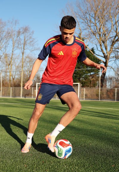 Joven futbolista masculino con el uniforme de España controlando un balón colorido en un campo de césped al aire libre bajo el sol.