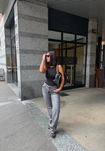 Cuerpo sin mangas negro, jeans rectos grises, tacones negros con tachuelas. Bolso negro con tachuelas. Superficie suave, entorno urbano.