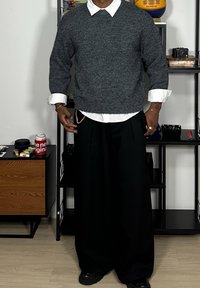 Maglione grigio lavorato a maglia con scollo rotondo sopra una camicia bianca con colletto. Pantaloni larghi neri e scarpe nere. Nota: accessori dorati visibili.
